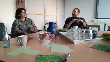 Zwei Personen sitzen an einem Tisch in einem Besprechungsraum. Auf dem Tisch befinden sich Getränke in Gläsern, eine Kanne, Geschirr und grüne Karten. Die Personen scheinen sich zu unterhalten und haben eine entspannte Atmosphäre.