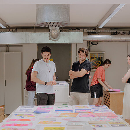 Design-Studierende betrachten sich Entwürfe auf einem Tisch am Campus unter den Eichen