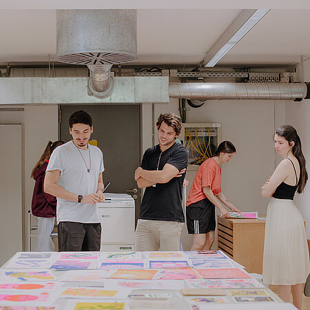 Design-Studierende betrachten sich Entwürfe auf einem Tisch am Campus unter den Eichen