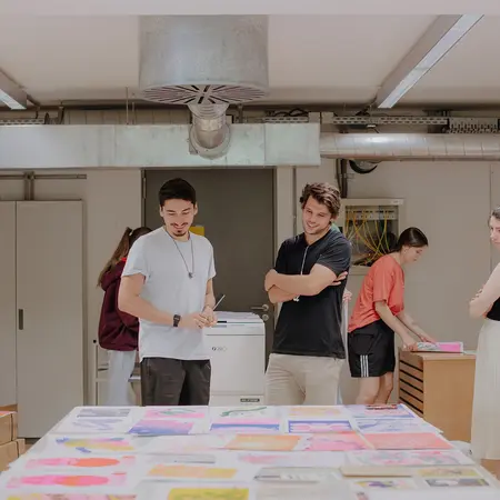 Design-Studierende betrachten sich Entwürfe auf einem Tisch am Campus unter den Eichen