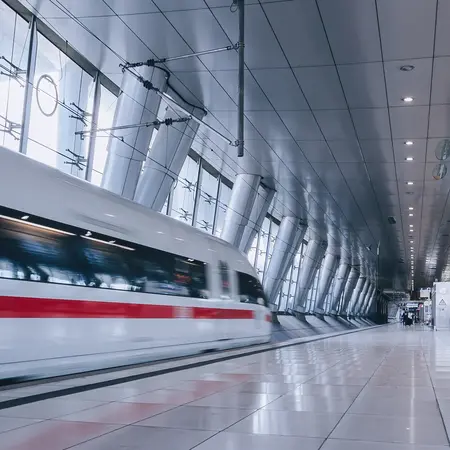 Ein moderner Bahnhof mit gläsernen Wänden und einem schnittigen Hochgeschwindigkeitszug, der vorbeifährt. Der Raum ist hell erleuchtet und zeigt eine klare, offene Architektur. Einige Reisende warten in der Ferne, während das Bahnsteigdesign klar und minimalistisch ist.
