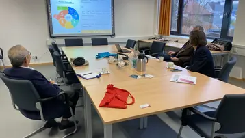 Eine Gruppe von Menschen sitzt an einem Konferenztisch und schaut auf ein Präsentationsbildschirm, der ein Farbschema und Diagramme zeigt. Materialien liegen auf dem Tisch, darunter ein rotes Stofftasche. Die Atmosphäre ist geschäftlich und konzentriert.