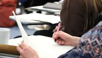 Eine Hand hält einen schwarzen Stift und schreibt auf einem Blatt Papier, während die andere Hand eine andere Seite hält. Im Hintergrund ist eine Tasse und ein Tisch sichtbar. Die Szene vermittelt eine Lern- oder Unterrichtssituation.
