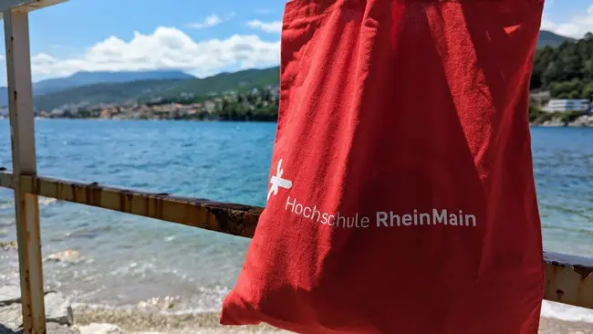 Auf dem Bild ist die rote Umhängetasche der HSRM zu sehen, die an einem Geländer hängt. Im Hintergrund ist das Meer zu sehen.