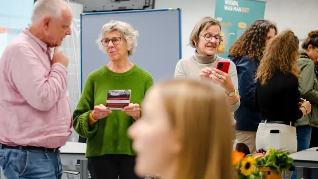 Eine Gruppe von Menschen steht in einem Raum und unterhält sich. Zwei Frauen halten Informationsmaterial und ein Smartphone. Eine Frau in der Mitte ist mit dem Rücken zur Kamera. Im Hintergrund sind weitere Personen zu sehen, die miteinander sprechen.