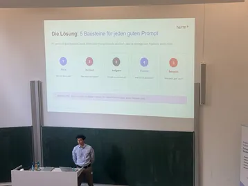 Ein Vortragender steht vor einer Präsentation, die die fünf Bausteine für einen guten Prompt beschreibt: Pacing, Kontext, Aufgabe, Format und Beispiel. Die Folie enthält auch das Logo der HSRM. Der Vortragende erklärt, wie diese Bausteine die Qualität von Ergebnissen verbessern können.