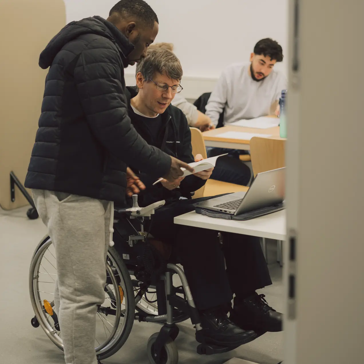 Zwei Männer diskutieren an einem Tisch. Einer sitzt in einem Rollstuhl und schaut auf ein Dokument, während der andere ihm etwas zeigt. Im Hintergrund ist ein dritter Mensch, der am Laptop arbeitet. Der Raum ist hell und modern gestaltet.