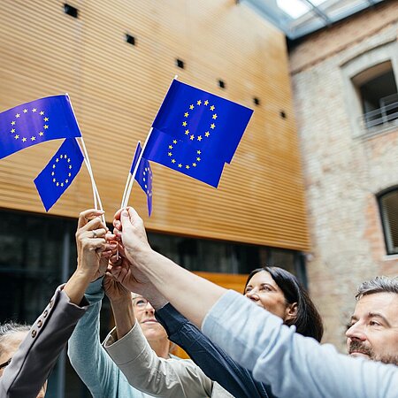 ERASMUS+ Mehrere Personen stehen in einem Kreis und halten kleine Europafraggen nach oben in die Mitte