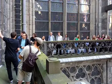 A group of architectural heritage students are standing on a balcony of Cologne Cathedral. Some are discussing, while others are listening attentively.