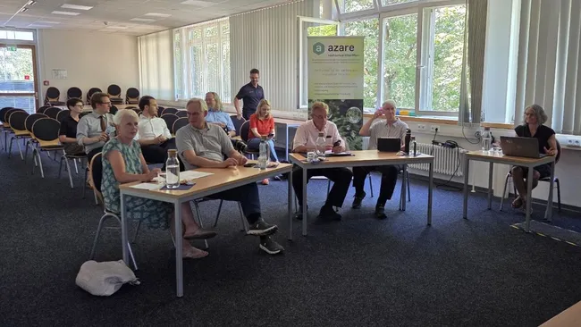 Eine Gruppe von Menschen sitzt an Tischen in einem Konferenzraum. Einige Personen nutzen Laptops, während andere miteinander sprechen. Im Hintergrund steht ein Banner mit der Aufschrift "azare". Die Umgebung wirkt hell und freundlich.
