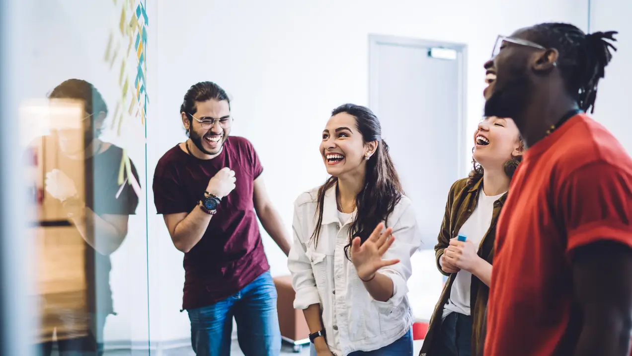 Eine Gruppe von vier Menschen lacht und hat Spaß in einem hellen Raum. Ihre freundlichen Gesichter zeigen Freude und konzentrierte Interaktion, während sie miteinander sprechen und lachen. Die Atmosphäre ist lebhaft und positiv, was die Gemeinschaft widerspiegelt.