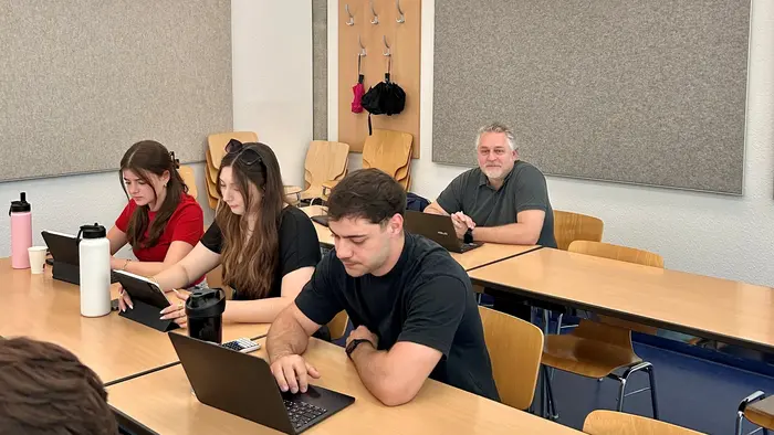 Im Bild sitzen mehrere Personen an einem Tisch in einem Klassenzimmer. Drei junge Erwachsene arbeiten konzentriert an Laptops, während eine ältere Person im Hintergrund sitzt und zuschaut. Die Umgebung ist hell und modern, mit einer grauen Wand im Hintergrund.