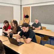 Im Bild sitzen mehrere Personen an einem Tisch in einem Klassenzimmer. Drei junge Erwachsene arbeiten konzentriert an Laptops, während eine ältere Person im Hintergrund sitzt und zuschaut. Die Umgebung ist hell und modern, mit einer grauen Wand im Hintergrund.