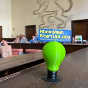 Auf einer Holzbank im Schwurgerichtsaal des Alten Gerichts in Wiesbaden steht der 3-D-gedruckte Pokal des Green Innovation Camps. Im Hintergrund ist an der Wand der hessische Löwe zu sehen.