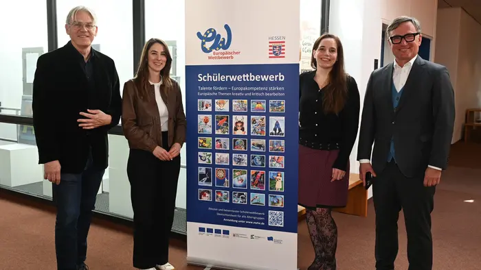 Eine Gruppe von vier Personen steht vor einem Banner, das Informationen zu einem Schülerwettbewerb zeigt. Die Personen lächeln und posieren in einer Innenraumumgebung. Das Banner enthält mehrere bunte Bilder und Text über den Wettbewerb.