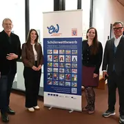 Eine Gruppe von vier Personen steht vor einem Banner, das Informationen zu einem Schülerwettbewerb zeigt. Die Personen lächeln und posieren in einer Innenraumumgebung. Das Banner enthält mehrere bunte Bilder und Text über den Wettbewerb.