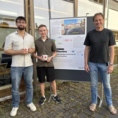 Drei Männer stehen vor einem Plakat, das ein Projekt namens "M@chine" präsentiert. Sie halten jeweils ein kleines Objekt in der Hand. Die Männer wirken, als würden sie stolz auf ihre Präsentation sein. Im Hintergrund sind Fenster und ein Teil des Gebäudes sichtbar.