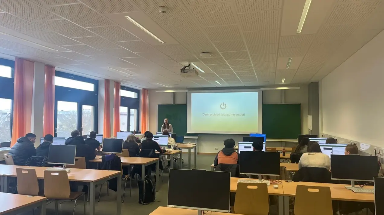 Ein Wohnzimmer ähnlicher Raum mit mehreren Computertischen. Vorne präsentiert eine Lehrerin an einem Bildschirm mit einem Power-Button-Symbol. Die Klasse besteht aus mehreren Schülern, die auf ihre Computer schauen, während sie der Lehrerin zuhören.