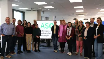 Eine Gruppe von 16 Personen steht in einem Büro. Sie lächeln und posieren vor einem Bildschirm, auf dem das Logo „ASO“ und der Text „Arbeitsgemeinschaft Soziale Arbeit in der Onkologie“ zu sehen ist. Der Raum ist hell und modern gestaltet.