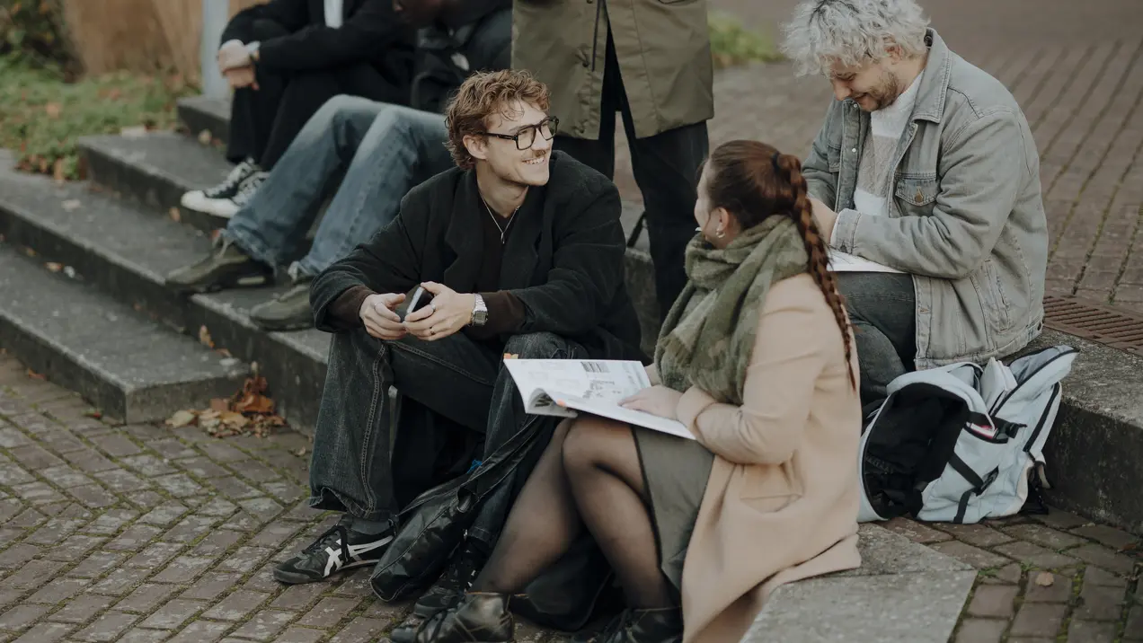 Eine Gruppe junger Menschen sitzt auf Stufen. Zwei Personen, eine Frau und ein Mann, sind im Gespräch, während die anderen in Ruhe zusammensitzen. Die Frau hat einen Skizzenblock in der Hand. Die Szene vermittelt eine freundliche, entspannte Atmosphäre im Freien.