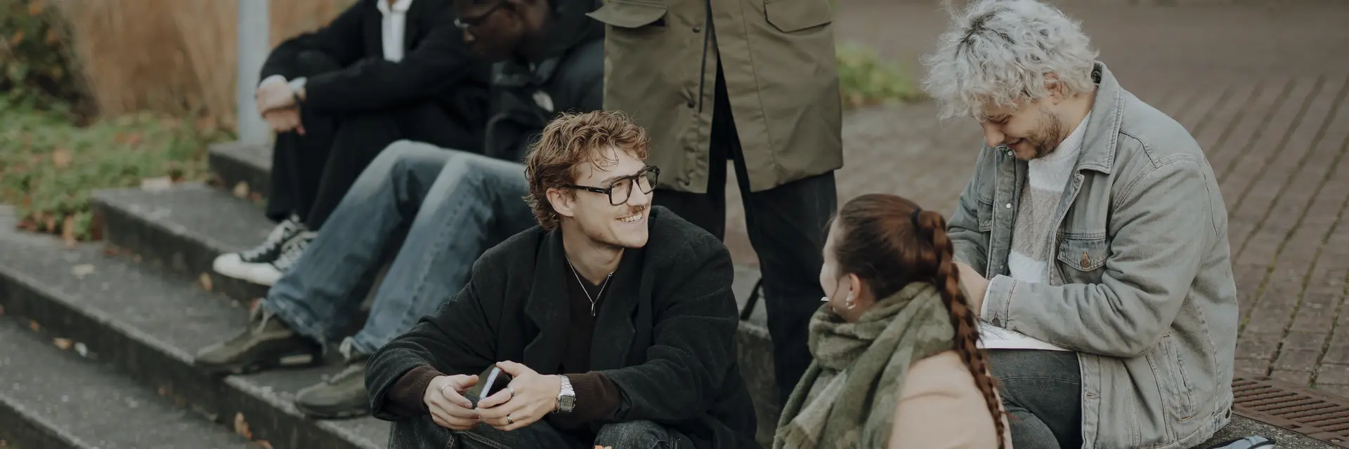 Eine Gruppe junger Menschen sitzt auf Stufen. Zwei Personen, eine Frau und ein Mann, sind im Gespräch, während die anderen in Ruhe zusammensitzen. Die Frau hat einen Skizzenblock in der Hand. Die Szene vermittelt eine freundliche, entspannte Atmosphäre im Freien.