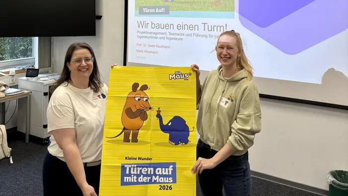 Zwei Personen halten ein gelbes Plakat mit dem Titel „Türen auf mit der Maus 2026“. Im Hintergrund sieht man einen Projektor mit Illustrationen. Die Atmosphäre ist freundlich und informativ, geeignet für eine Veranstaltung oder Präsentation.
