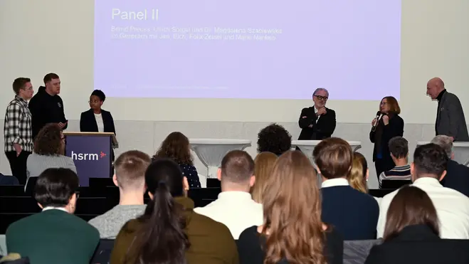 Bernd Preuss, Ulrich Sörgel, Dr. Magdalena Szablewska, Jan Eich, Felix Zeisel und Marie Nankep stehen auf der Bühne der Hochschule RheinMain, während eine Moderation stattfindet. Ein Bildschirm im Hintergrund zeigt den Text "Panel II". Die Teilnehmenden diskutieren verschiedene Themen beim 2. Wiesbadener Tag für sozial nachhaltiges Investieren.