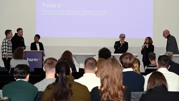 Bernd Preuss, Ulrich Sörgel, Dr. Magdalena Szablewska, Jan Eich, Felix Zeisel und Marie Nankep stehen auf der Bühne der Hochschule RheinMain, während eine Moderation stattfindet. Ein Bildschirm im Hintergrund zeigt den Text "Panel II". Die Teilnehmenden diskutieren verschiedene Themen beim 2. Wiesbadener Tag für sozial nachhaltiges Investieren.