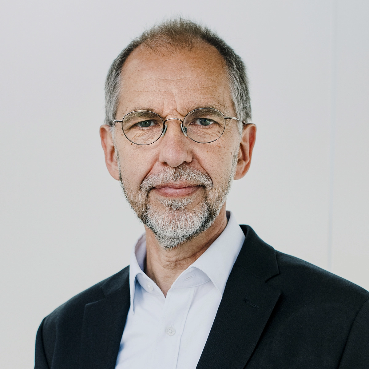 Ein älterer Mann mit Brille und grauem Bart trägt einen dunklen Anzug und ein weißes Hemd. Er schaut direkt in die Kamera und hat einen freundlichen Ausdruck. Der Hintergrund ist hell und neutral.