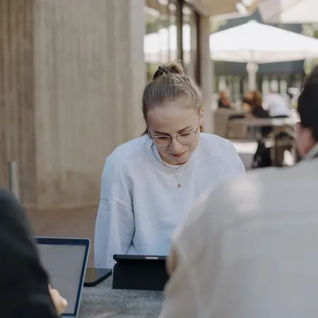 Eine junge Frau sitzt an einem Tisch im Freien und lächelt, während sie auf ein Tablet schaut. Zwei Männer, die sich ihr gegenüber befinden, sind von hinten zu sehen und arbeiten an Laptops. Um sie herum sind Sonnenblenden und eine entspannte Atmosphäre.