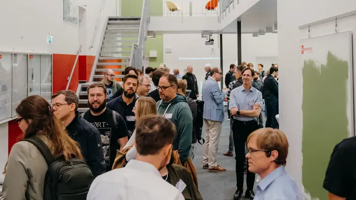 Eine Gruppe von Menschen steht in einem langen Flur, der auf einer Veranstaltung oder Konferenz ist. Einige Teilnehmer unterhalten sich, während im Hintergrund eine Treppe und Wände in verschiedenen Farben sichtbar sind. Die Atmosphäre wirkt gesellig und informell.