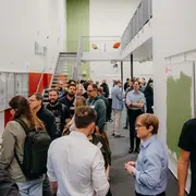Eine Gruppe von Menschen steht in einem langen Flur, der auf einer Veranstaltung oder Konferenz ist. Einige Teilnehmer unterhalten sich, während im Hintergrund eine Treppe und Wände in verschiedenen Farben sichtbar sind. Die Atmosphäre wirkt gesellig und informell.