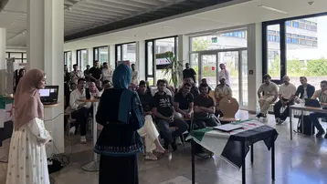 Eine Frau steht vor einer Gruppe von Menschen und präsentiert. Die Zuhörer sind aufmerksam und sitzen an Tischen, während eine Flagge im Vordergrund zu sehen ist. Der Raum ist hell erleuchtet, mit großen Fenstern, die einen Blick nach draußen ermöglichen.