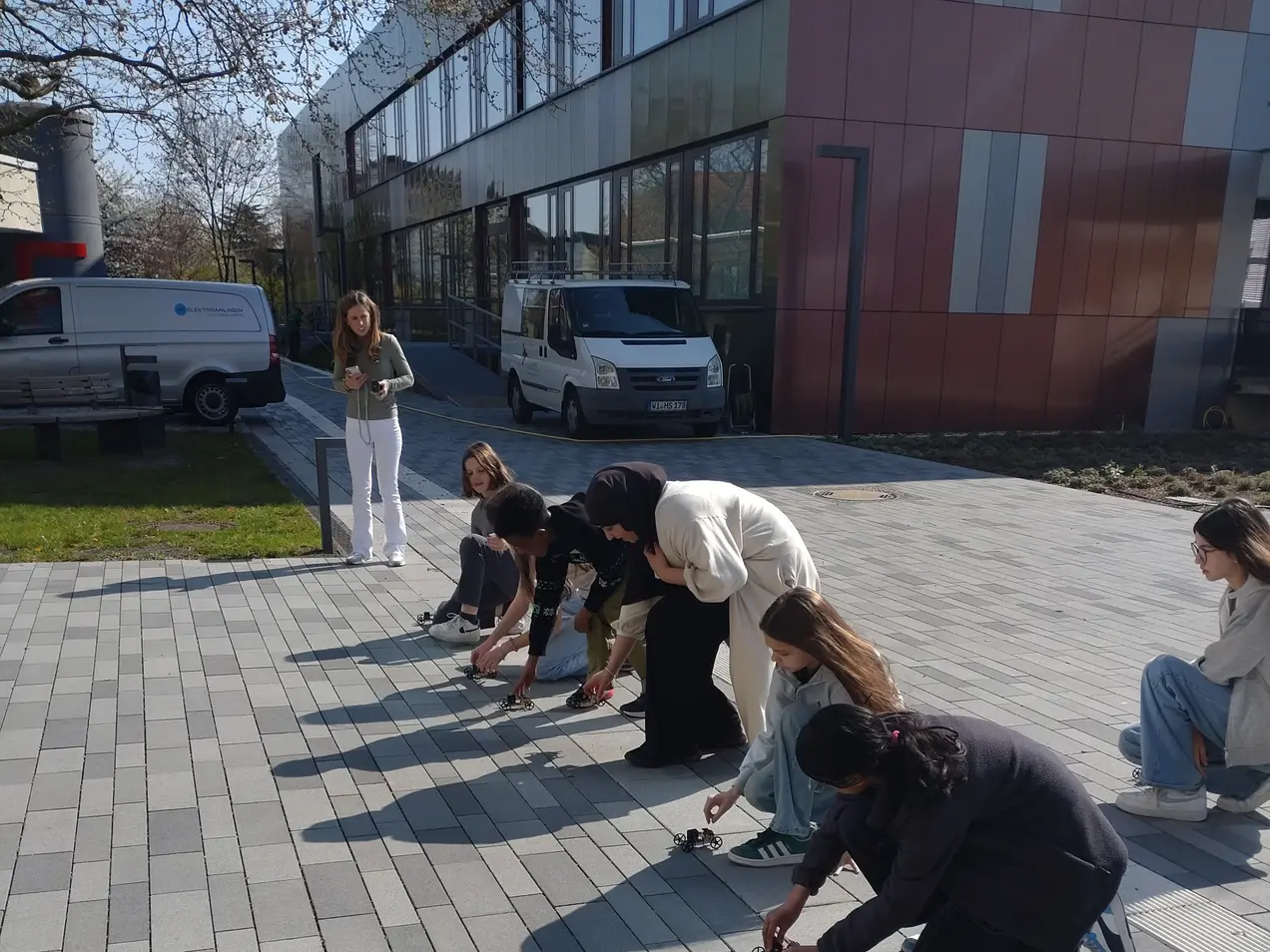 Eine Gruppe von sieben Personen steht und kniet auf einem gepflasterten Platz. Sie bücken sich und schauen konzentriert auf den Boden, als ob sie etwas untersuchen oder sammeln. Im Hintergrund sind ein Gebäude und zwei Fahrzeuge zu sehen.