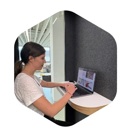 Eine junge Frau sitzt an einem Arbeitsplatz und tippt auf ihrem Laptop. Sie hat lange, braune Haare und trägt ein einfaches, weißes T-Shirt. Die Umgebung ist modern und hell. Im Hintergrund sind Teile des Büros sichtbar.