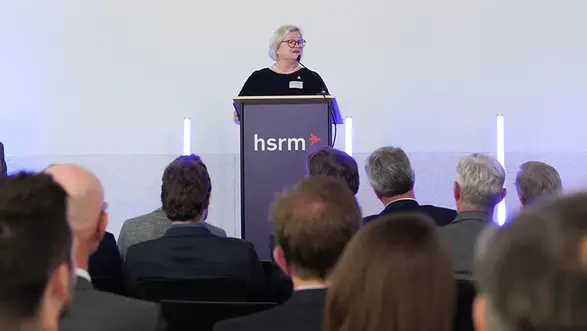 Prof. Dr. Eva Waller steht an einem Rednerpult und hält eine Ansprache. Im Hintergrund sind eine Menge Zuhörer in Anzügen zu sehen. Das Pult trägt das Logo "hsrm". Die Umgebung ist modern und hell.