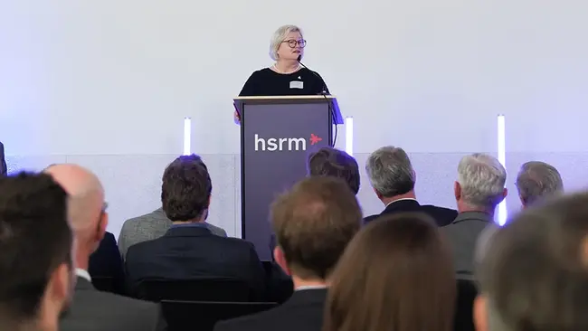 Prof. Dr. Eva Waller steht an einem Rednerpult und hält eine Ansprache. Im Hintergrund sind eine Menge Zuhörer in Anzügen zu sehen. Das Pult trägt das Logo "hsrm". Die Umgebung ist modern und hell.