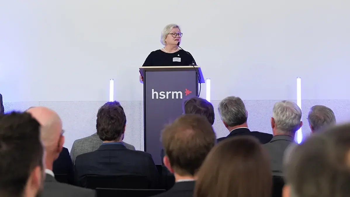 Prof. Dr. Eva Waller steht an einem Rednerpult und hält eine Ansprache. Im Hintergrund sind eine Menge Zuhörer in Anzügen zu sehen. Das Pult trägt das Logo "hsrm". Die Umgebung ist modern und hell.