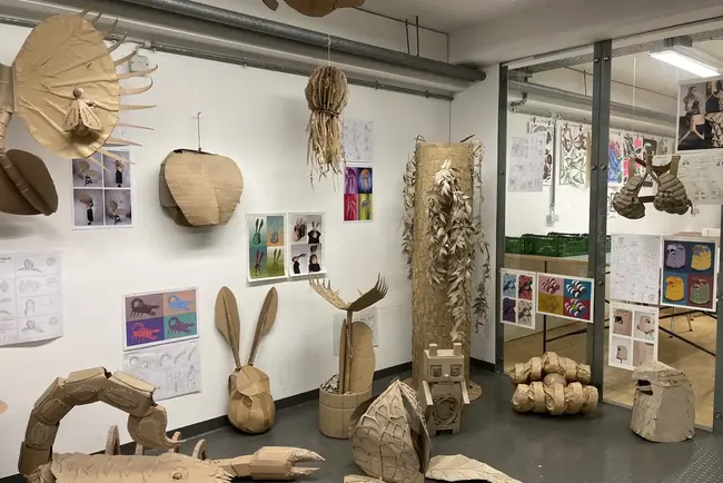 Der Raum zeigt eine kreative Ausstellung aus Pappmaché, die verschiedene Tierfiguren und -skulpturen darstellt, wie zum Beispiel einen Skorpion, einen Hasen und eine Schildkröte. Im Hintergrund hängen Skizzen und Illustrationen von Tieren an der Wand.