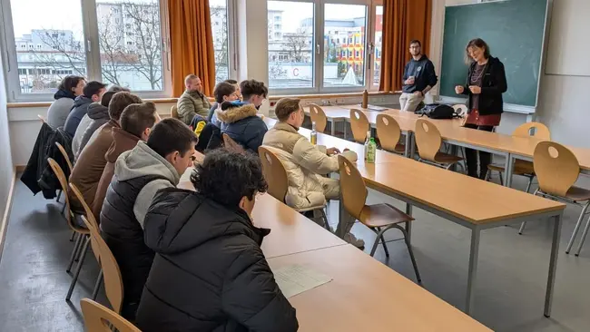 In einem Seminarraum sitzen mehrere Schülerinnen und Schüler an Tischen und hören zwei Lehrkräfte zu, die vorne sprechen. Die Fenster zeigen den Campus Rüsselsheim im Hintergrund. Die Atmosphäre wirkt aufmerksam und lehrreich.