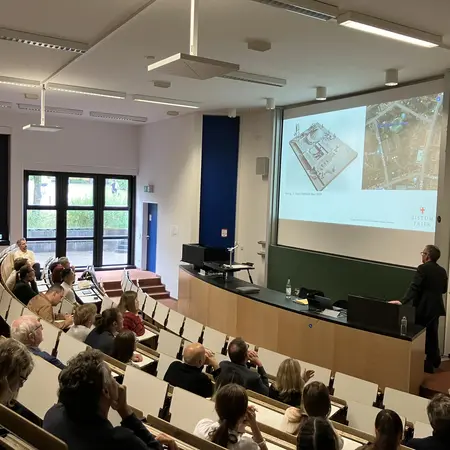 Eine Präsentation anlässlich der Gespräche zur Baukultur in einem Seminarraum an der HSRM. Ein Referent vor einem Publikum steht. An der Leinwand ist eine technische Zeichnung und ein Bild einer Baustelle zu sehen. Die Teilnehmer sitzen in Reihen und hören aufmerksam zu.