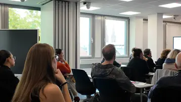 Eine Gruppe von Menschen sitzt in einem modernen Schulungsraum. Einige Teilnehmer hören aufmerksam zu, während ein Referent präsentiert. Im Hintergrund sind Fenster mit Tageslicht und eine Präsentationsleinwand sichtbar. Die Atmosphäre wirkt konzentriert und informativ.