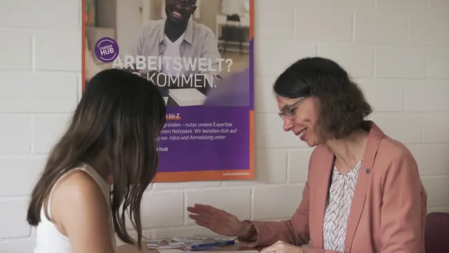 Zwei Personen sitzen an einem Tisch. Eine Frau mit langen Haaren und eine ältere Frau im rosa Blazer unterhalten sich. Im Hintergrund hängt ein Plakat mit dem Text „ARBEITSWELT? KOMMEN.“ Die Atmosphäre wirkt freundlich und unterstützend.