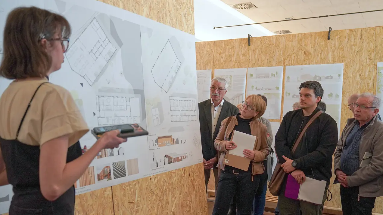 A presentation displays various architectural plans on a wall. A student stands at the front and explains her design, while a group of four people listen attentively.