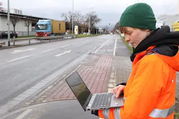 Eine Person in einer orangefarbenen Warnjacke und grüner Mütze steht vor einer Straße und arbeitet mit einem Laptop. Im Hintergrund fährt ein Lkw. Der Himmel ist bewölkt und die Umgebung wirkt städtisch.