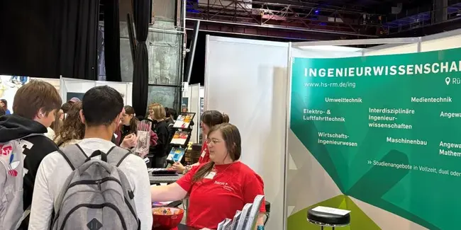 Eine Messe für Ingenieurwissenschaften mit einem Informationsstand. Eine Person in rotem Shirt erklärt Besucherinnen und Besuchern Angebote und Studiengänge. Im Hintergrund sind weitere Menschen zu sehen, die sich informieren. Broschüren und ein Wasserbehälter sind auf dem Tisch platziert.