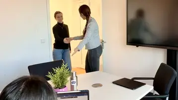 Ein Mann und eine Frau stehen sich in einem Büro gegenüber und schütteln sich die Hände. Im Vordergrund ist ein Tisch mit einem Laptop und einer Pflanze zu sehen. Der Raum hat eine helle und freundliche Atmosphäre.