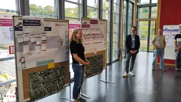 Eine Präsentation findet in einem lichtdurchfluteten Raum statt, mit mehreren Flipcharts und einem großen Stadtplan im Hintergrund. Eine Frau steht und erklärt, während zwei Männer und eine weitere Person aufmerksam zuhören. Die Atmosphäre ist informativ und interaktiv.