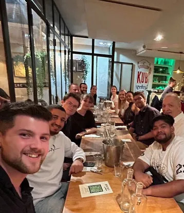 Eine Gruppe von etwa 15 Personen sitzt an einem langen Tisch in einem Restaurant. Die Atmosphäre ist freundlich und gesellig, mit Getränken auf dem Tisch. Einige Personen lächeln direkt in die Kamera, während andere entspannt miteinander sprechen.