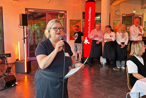 Eine Frau in Kellnerschürze spricht in ein Mikrofon.